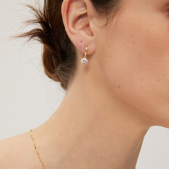 Close-up of a person wearing gold hoop earrings with a clear gemstone on a neutral background