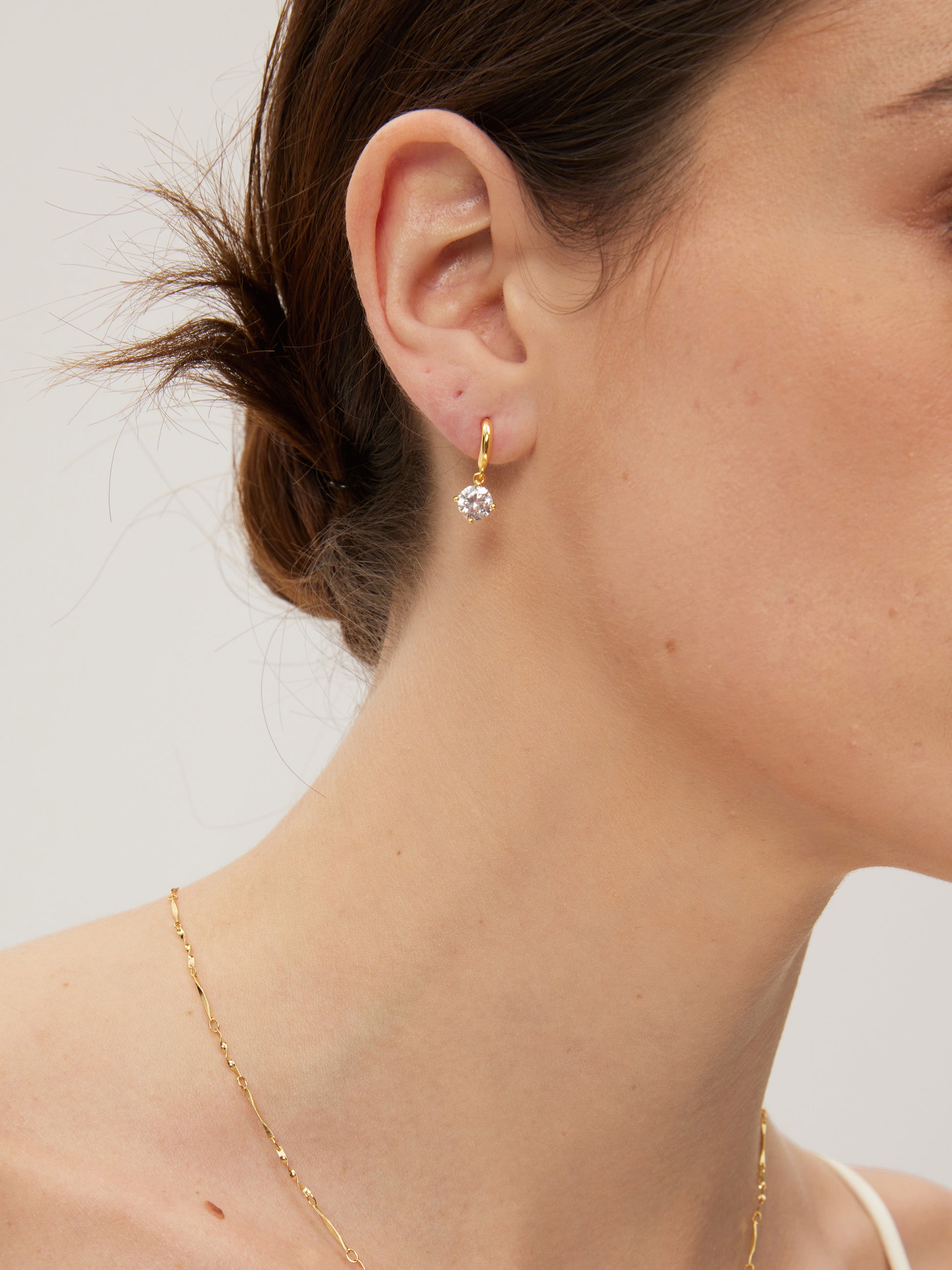 Close-up of a person wearing gold hoop earrings with a clear gemstone on a neutral background