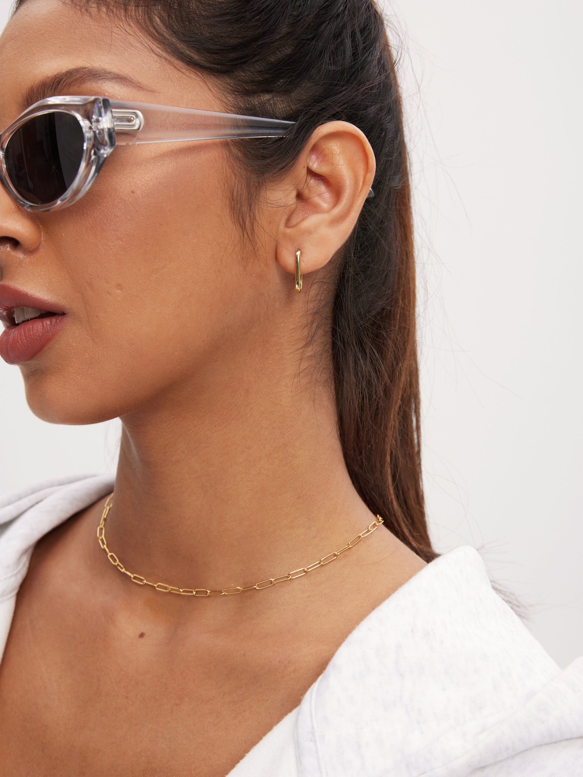 Woman wearing clear sunglasses, gold hoop earrings, and a gold chain necklace on a white background