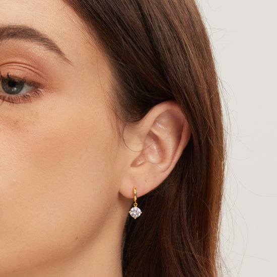 Close-up of a person wearing a gold earring with a clear gemstone on a neutral background