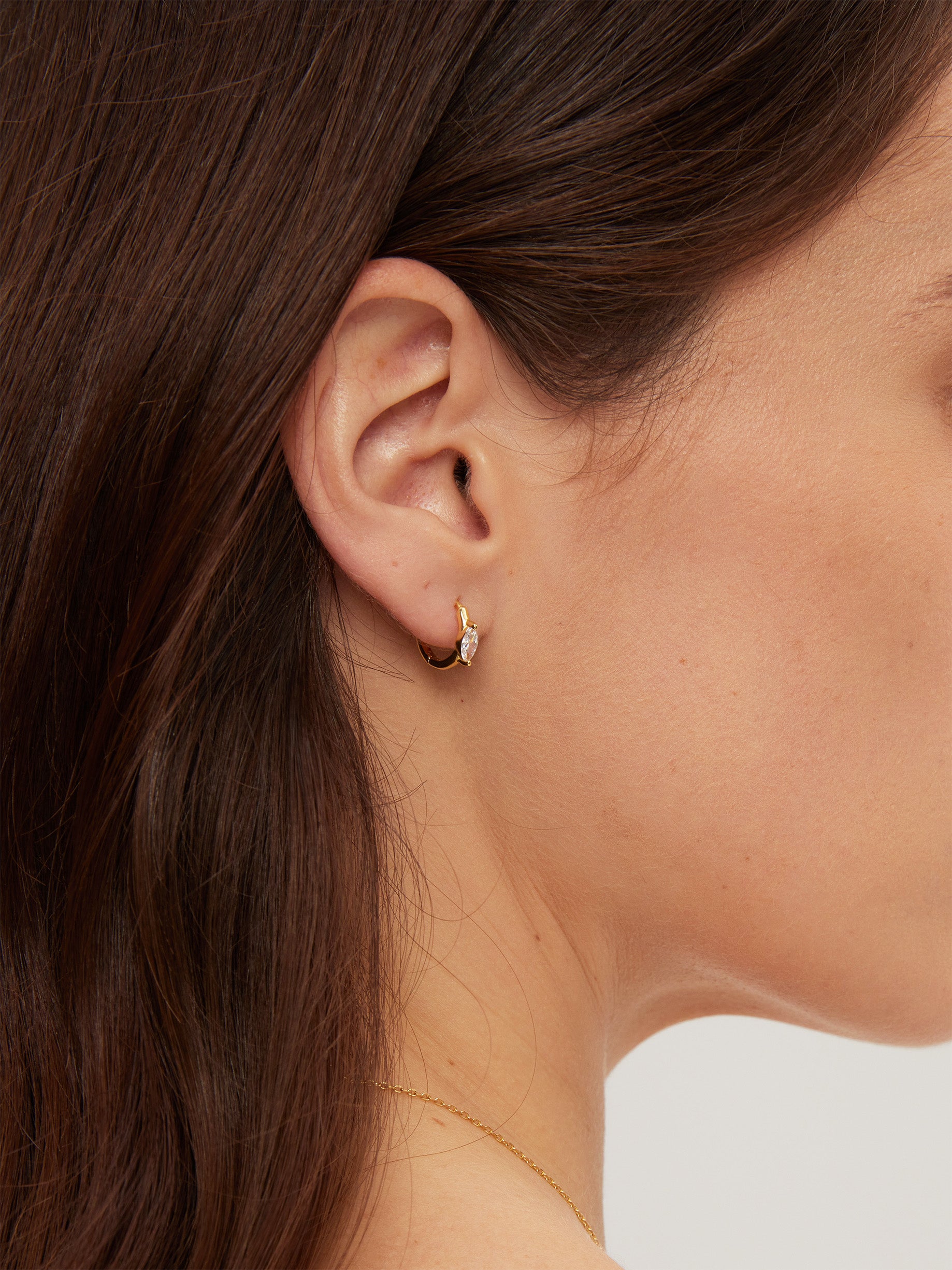 Close-up of an ear wearing a gold earring with a clear gemstone on a neutral background
