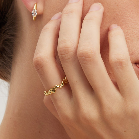 Close-up of a hand wearing a gold molten ring with an earring in the background.