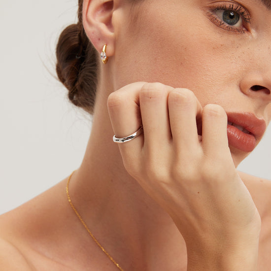 Close-up of a woman wearing gold earrings, silver classic ring, and necklace on a neutral background