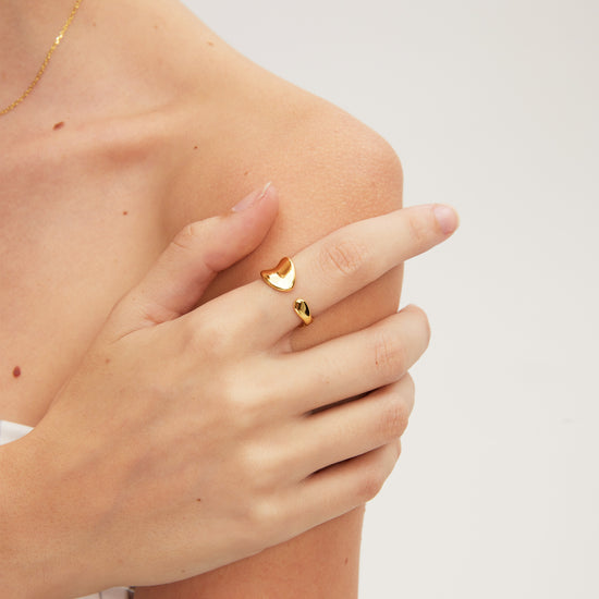 Gold ring with a heart design worn on a finger against a neutral background